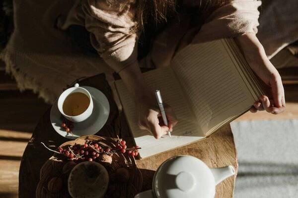 Person writing in a notebook beside a laptop and a mug